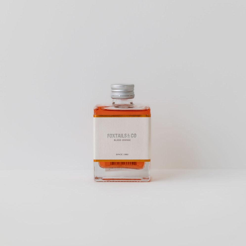 A small, clear glass bottle filled with orange liquid, labeled "Foxtails & Co. Blood Orange Aperitivo," against a plain white background.