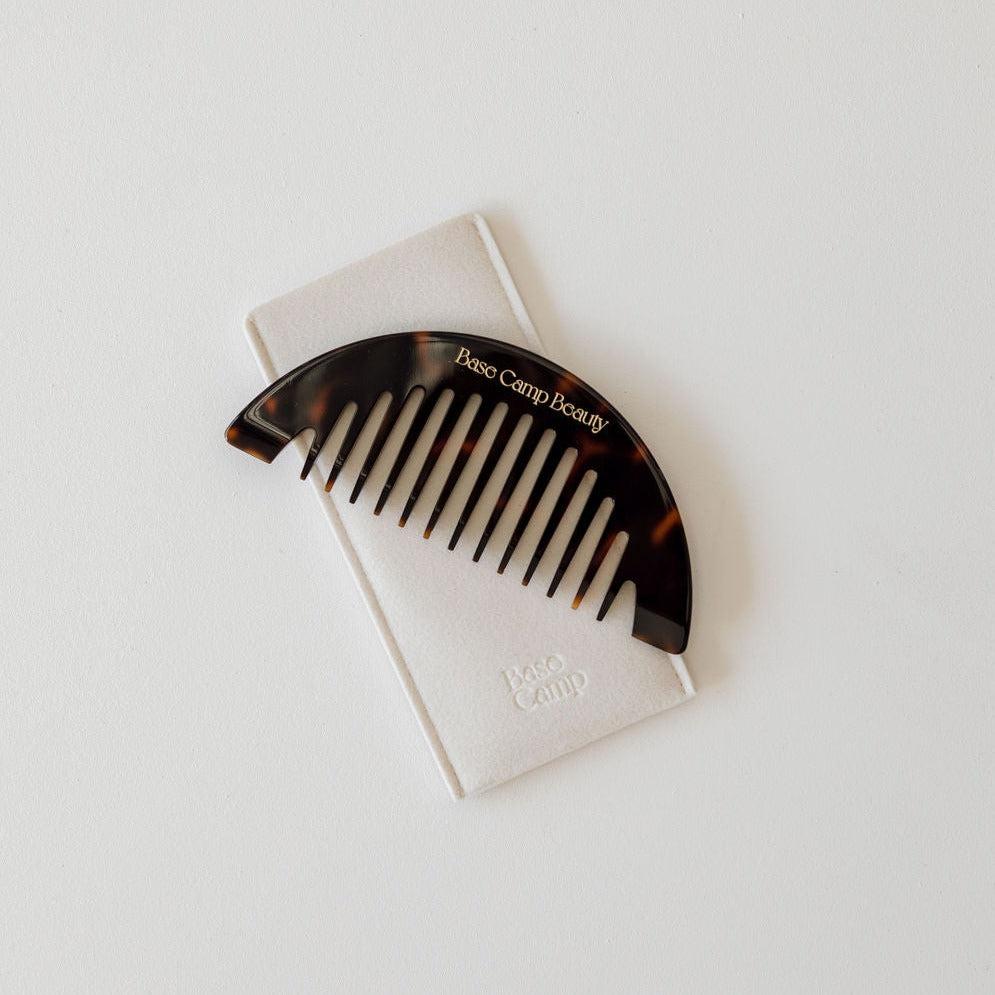 A tortoise wide-tooth crescent comb with a white label on it, ideal for hair care and hair therapy by Base Camp Beauty.