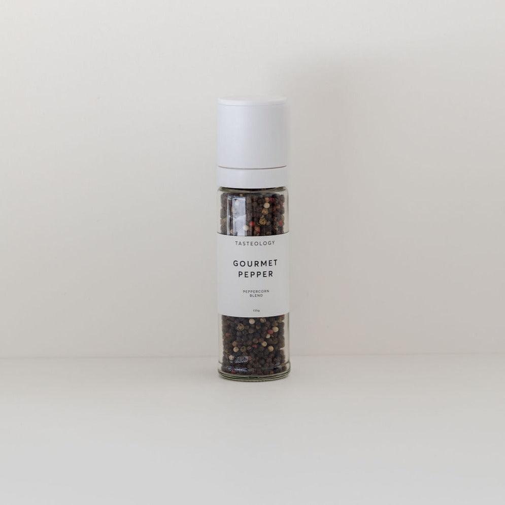 A clear glass grinder bottle containing assorted Tasteology Gourmet Pepper labeled on a plain beige background.