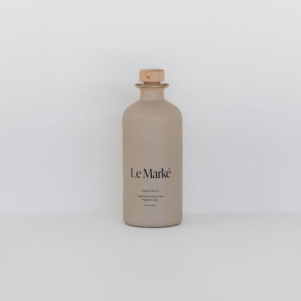 A beige ceramic bottle with a cork lid labeled "Le Marké olive oil" against an ivory background.