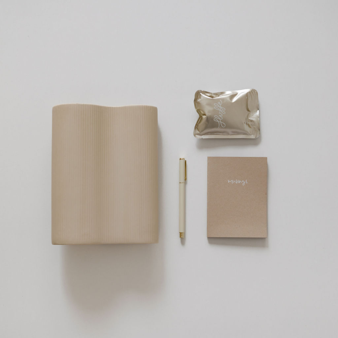 A "biglittlegifting" beige notebook and a pen on a white surface.