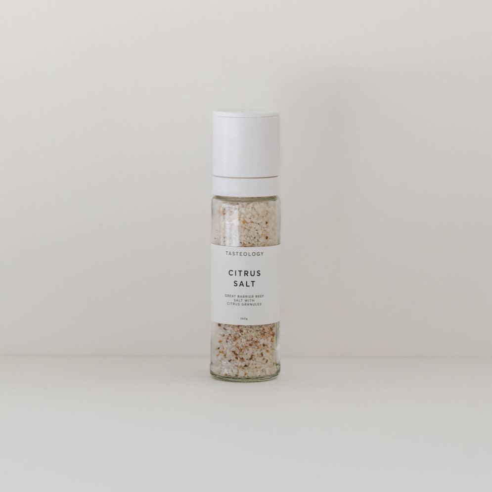 Clear glass bottle of Tasteology citrus salt labeled "Tasteology" for enhancing seafood and fish dishes, against a plain, light beige background.
