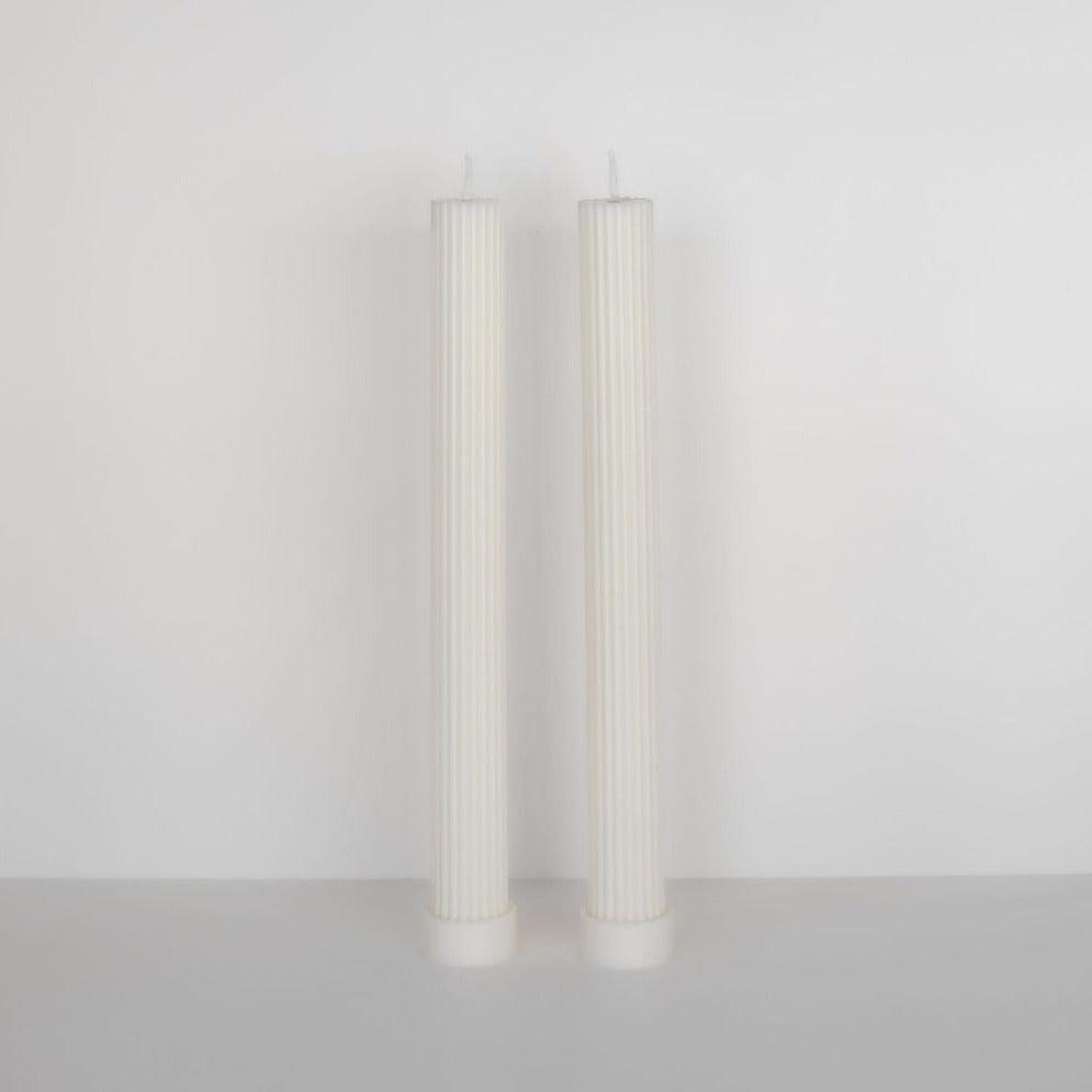 Two tall, Black Blaze column pillar candle duo in white standing upright against a plain gray background.