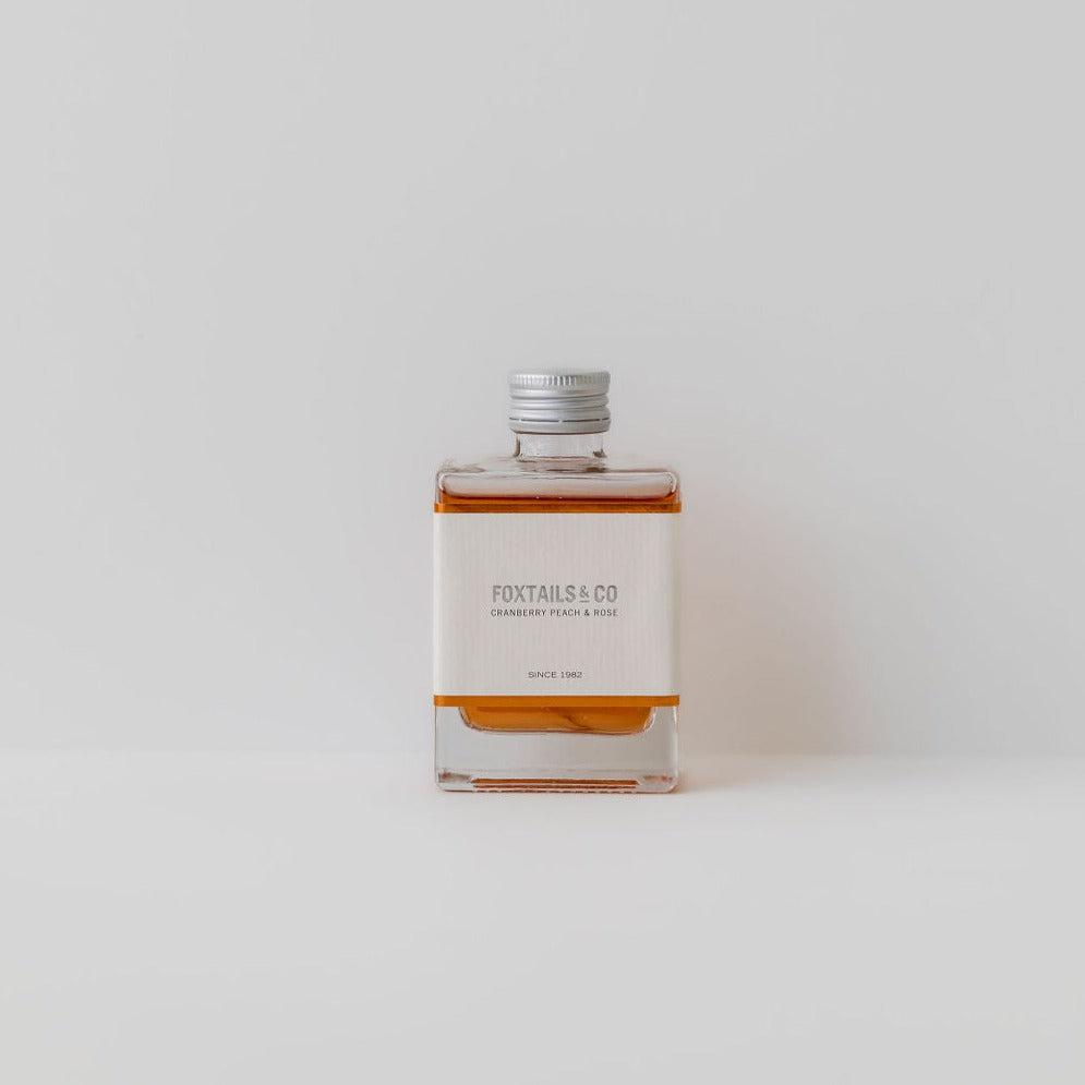 A bottle of Foxtails & Co cranberry peach & rose sitting on a white surface, surrounded by peaches and cranberry.
