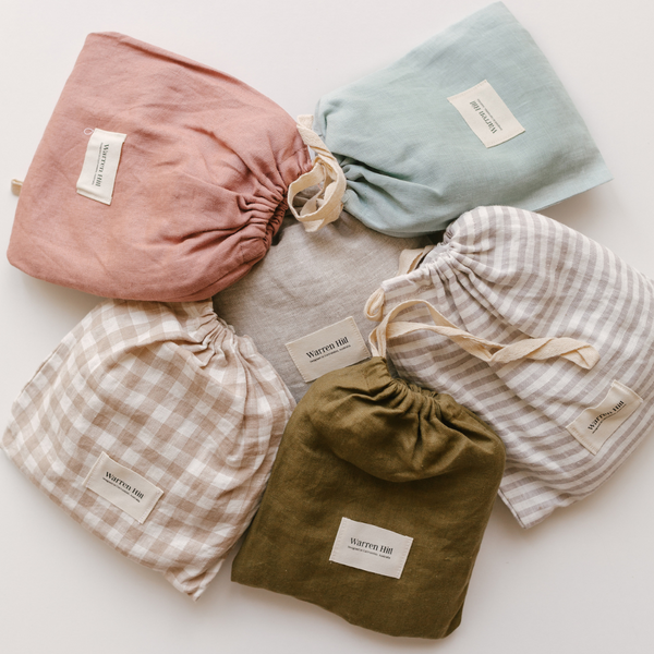 Assorted drawstring bags in different colors and patterns including pink, teal, grey, plaid, and olive, labeled with tags on a Warren Hill french linen fitted cot sheet in beige gingham.