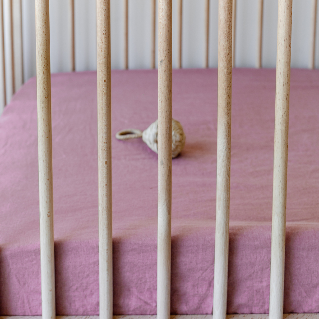 A baby crib with a Warren Hill French Linen Fitted Cot Sheet in Smoke Pink.