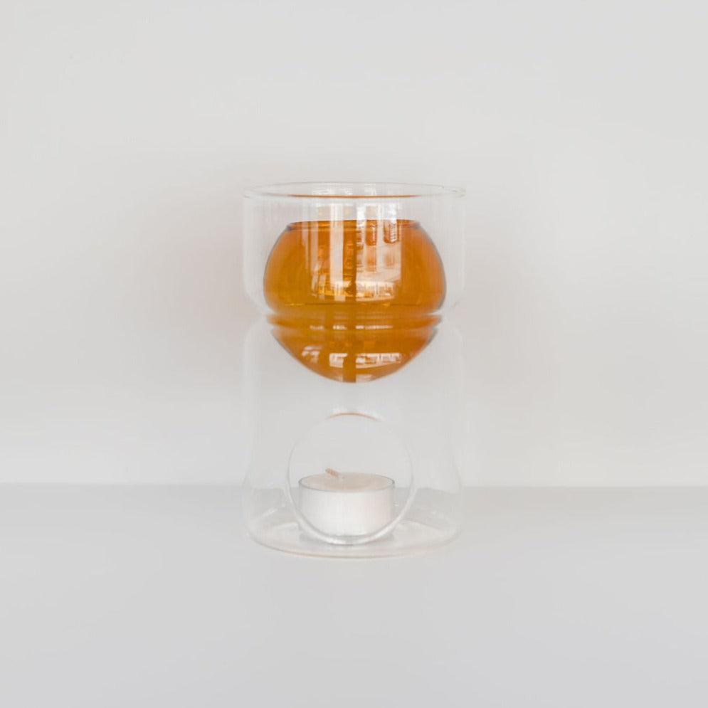 A Fazeek clear glass oil burner with a lit soy tealight candle underneath and amber-colored essential oil above, set against a white background.