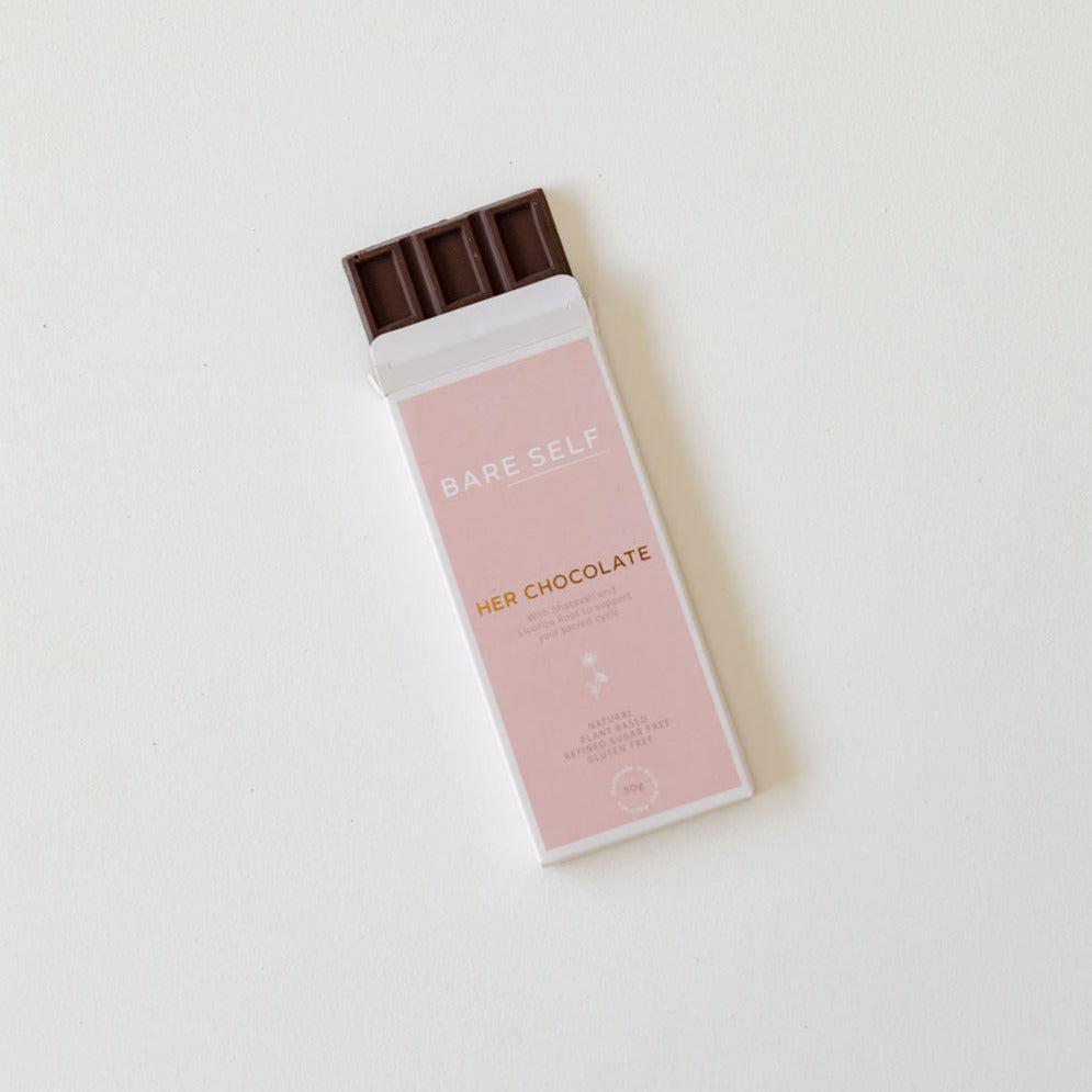 A pink Bare Self chocolate bar, named "Her Chocolate," rests elegantly on a pristine white surface. This delectable treat not only satisfies your sweet tooth but also promotes women's reproductive health.