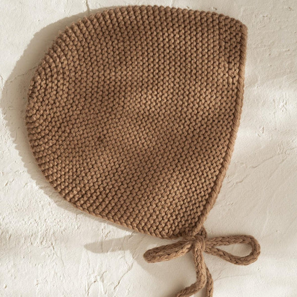 An Illoura the Label organic cotton knitted illoura baby bonnet in chocolate on a white wall.