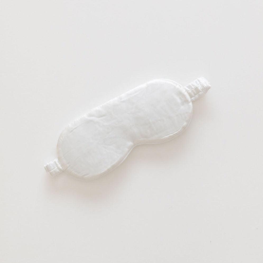 A pearl white, padded silk eye mask from The Silk Collection lying on a plain, light background.