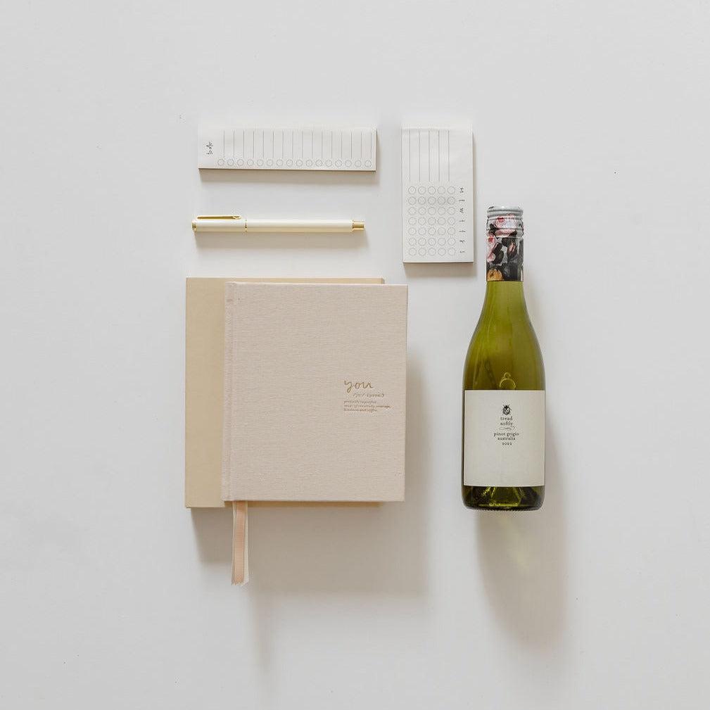 A promotion celebration with a bottle of BigLittleGifting's You Time Plus Wine and a notebook on a white surface.