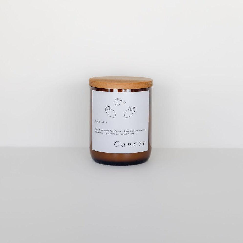 A Zodiac Candle in a glass jar with a wooden lid, labeled "Cancer" featuring a crab illustration from our Zodiac Candle Range by The Commonfolk Collective, placed against a plain white background.