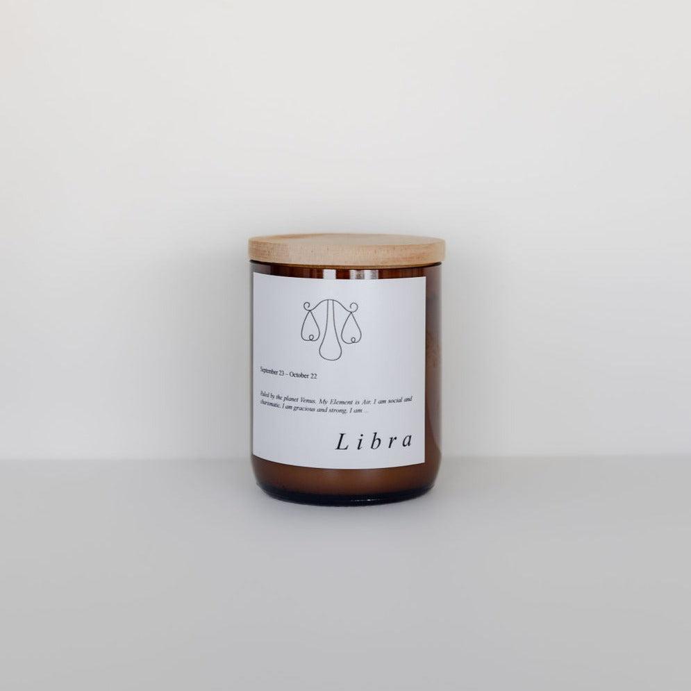 A The Commonfolk Collective Zodiac Candle Range soy candle wax in a clear jar with a wooden lid labeled "Libra", featuring a scale graphic and text, on a white background.