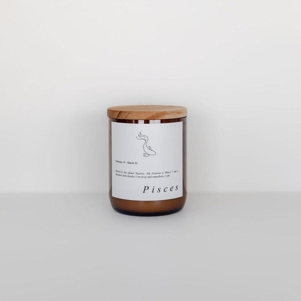 A zodiac candle in a clear glass jar with a wooden lid, labeled 'Pisces' and featuring a line drawing of a fish, placed against a plain white background by The Commonfolk Collective.