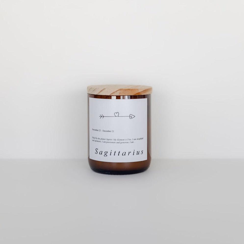 A Commonfolk Collective candle labeled "Sagittarius" with a wooden lid against a white background, featuring simple graphic symbols and minimalist text.