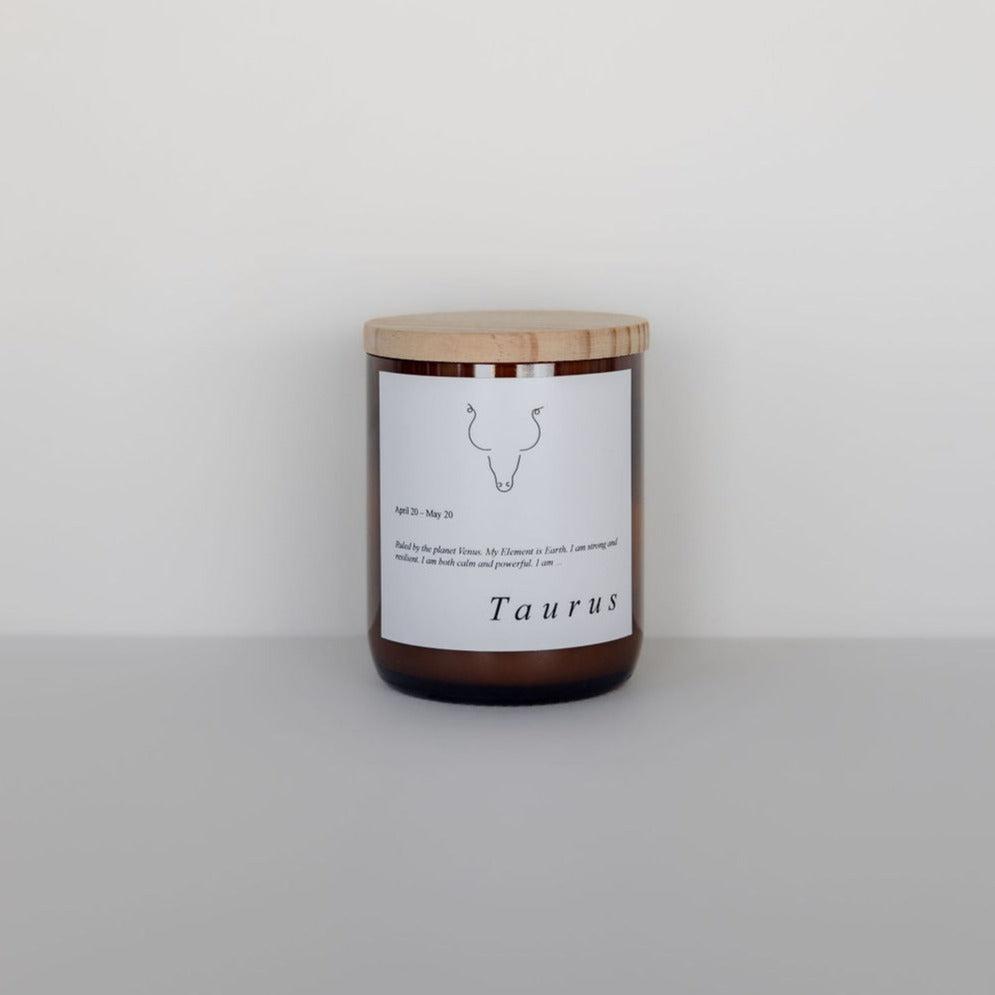 A The Commonfolk Collective Zodiac candle labeled "Taurus" with a simple bull icon, wooden lid, and text detailing scent notes, against a light gray background.